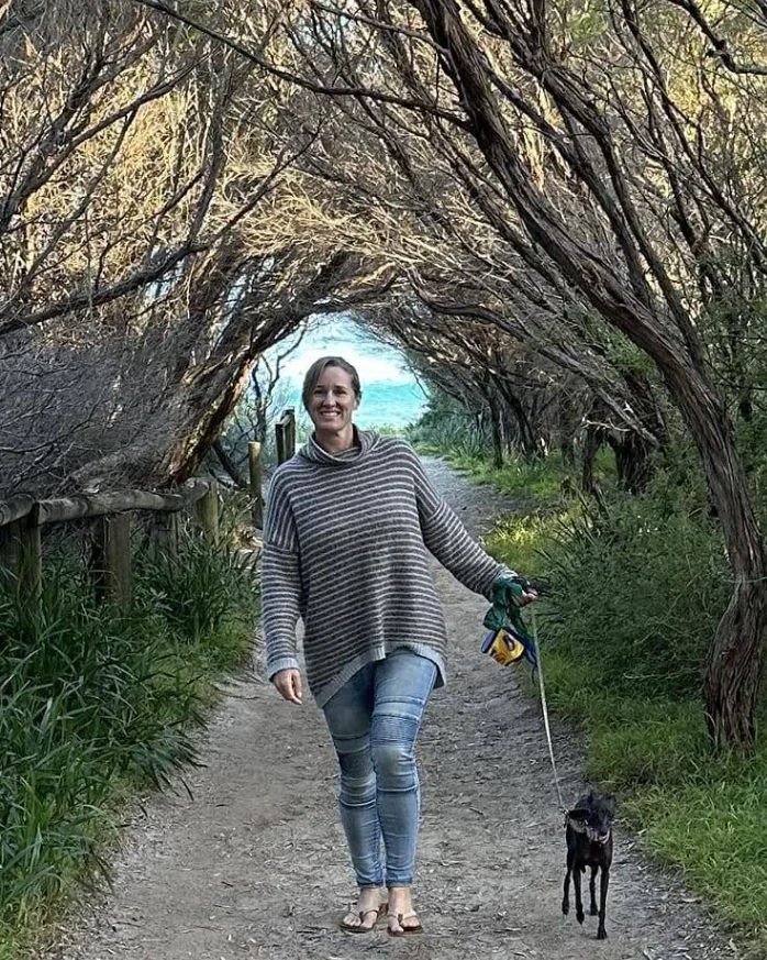 Kris smiling as she walks her small black dog along a sandy path, through a tunnel of coastal tea-trees opening onto the ocean.