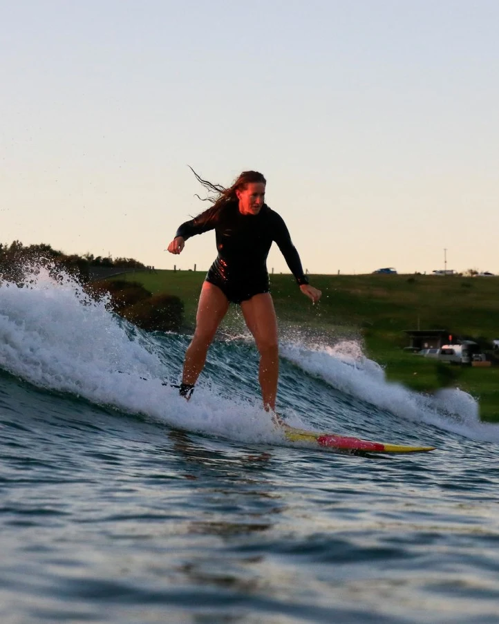 Kris riding a wave toward the camera at dusk, hair flying, a grassy headland behind her.