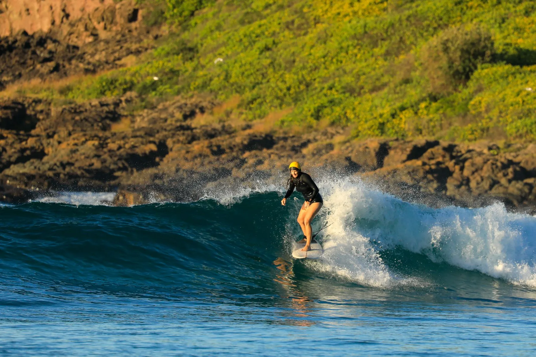 Kris in her yellow helmet riding a curling blue wave in front of a rocky, green headland.