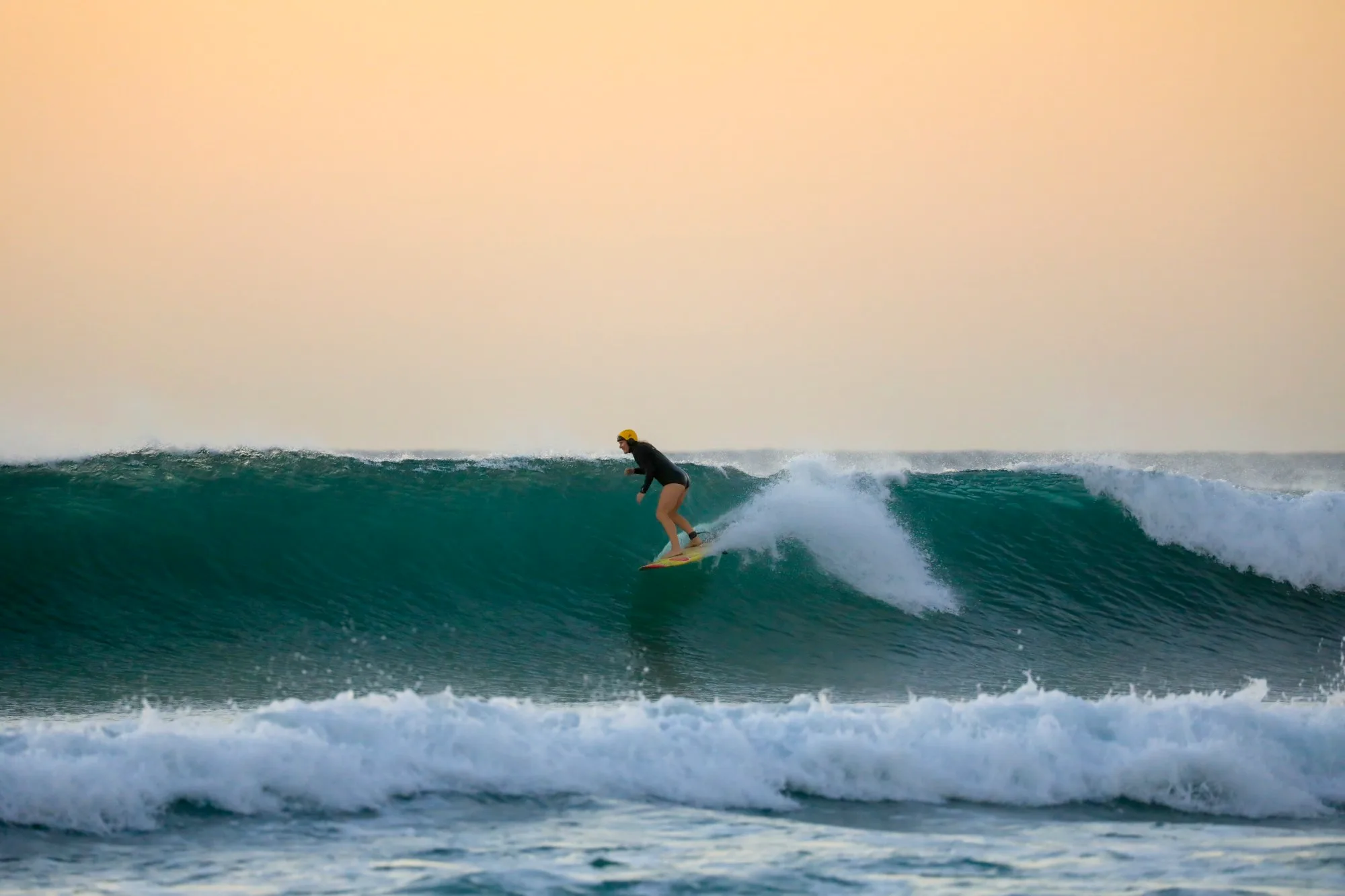Kris in her yellow helmet riding a green wave under an apricot sunset sky.